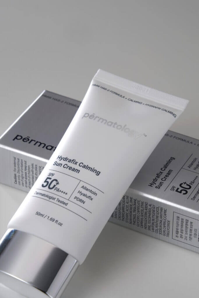 Tube of sunscreen labeled "SPF 50+" resting on its box at a slight angle on a light grey background; the white tube shows product text and a silver cap visible at the base of the box.