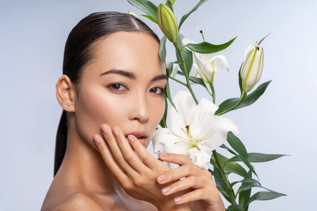 Close-up of a woman with clear skin gently touching her face, white lilies in soft focus behind her.